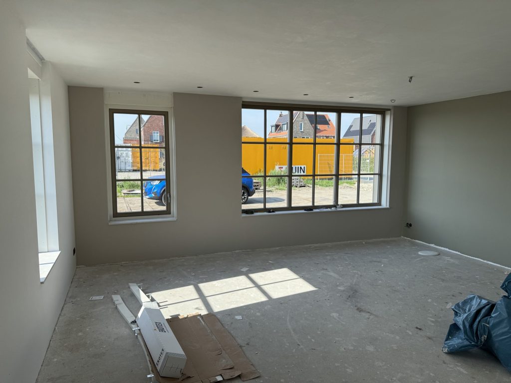 Lege nieuwbouwkamer met groengrijze wanden en grote roedenramen die uitkijken op de bouwplaats