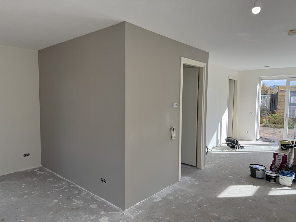 Open benedenverdieping van een nieuwbouwhuis met een taupe wand, betonvloer en emmers verf bij de tuindeur