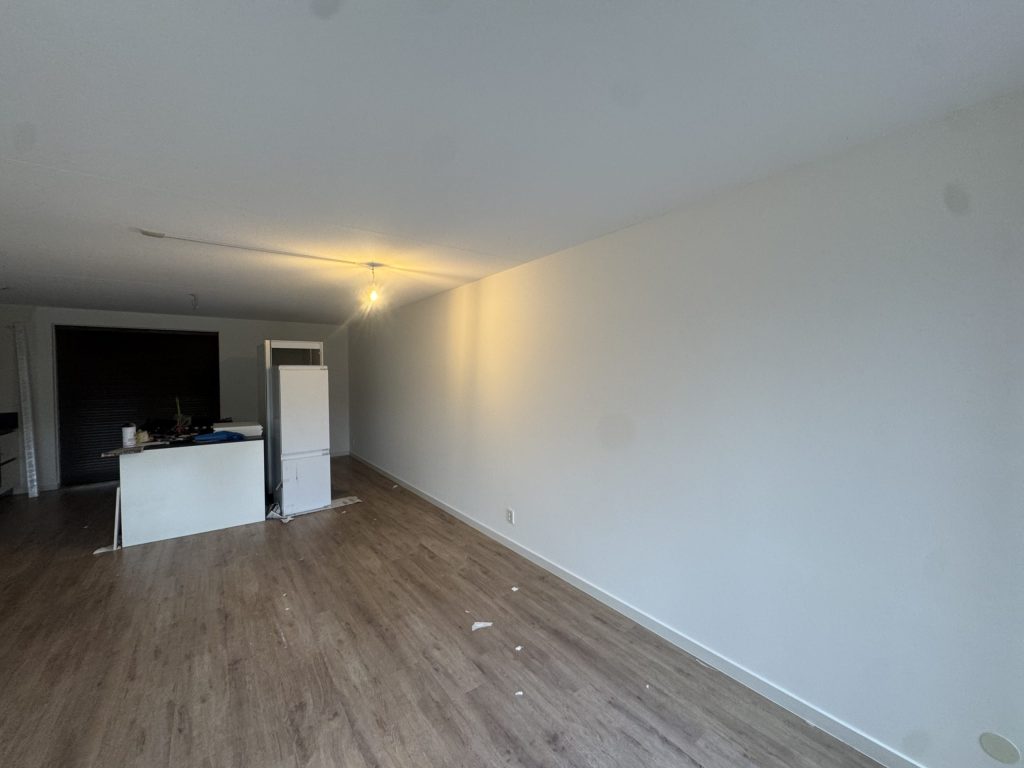 Lange wand van een nieuw appartement, strak wit afgewerkt boven een houten vloer met de keuken achterin